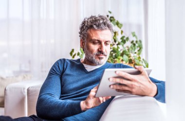 Evde orta yaşlı, çekici bir adamın portresi. Tablette TV programı izliyor, oturma odasında kanepede oturuyor, gülümsüyor..