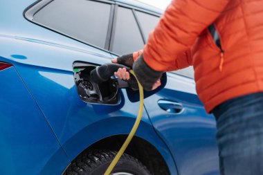Soğuk karlı bir günde adam elektrikli arabayı şarj ediyor. Şarj aletini şarj limanına koyan adama yaklaş. Kış mevsiminde elektrikli araçların şarj edilmesi ve kullanılması