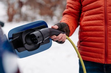 Soğuk karlı bir günde adam elektrikli arabayı şarj ediyor. Şarj aletini şarj limanına koyan adama yaklaş. Kış mevsiminde elektrikli araçların şarj edilmesi ve kullanılması