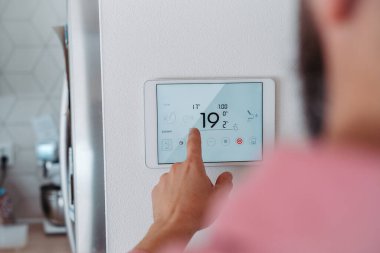 Man using smart thermostat, adjusting or lowering heating temperatures at his home. Concept of sustainable, efficient, and smart technology in home heating and thermostats. clipart