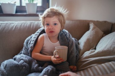 Küçük çocuk fincandan çay içiyor. Hasta çocuk kanepede sıcak battaniyenin altında oturup bitki çayı içiyor.