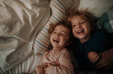 Candid portrait of siblings lying together in bed, looking at camera and laughing. Top view. clipart