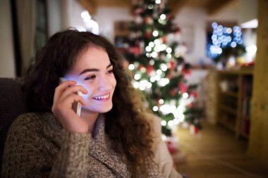 Genç bir kadın yılbaşı telefonu ediyor, oturma odasında Noel süsleriyle oturuyor. Noel atmosferi.