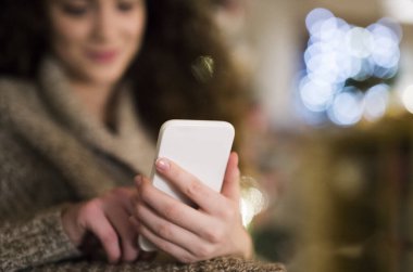 Elinde akıllı telefon tutan ve yılbaşı mesajı okuyan genç bir kadın. Oturma odasında Noel süsleriyle oturuyor..
