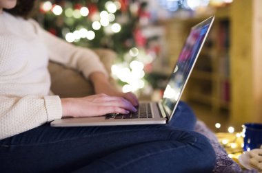 Young woman is shopping for Christmas gifts online using her laptop, sitting on couch in living room with Christmas decorations. clipart