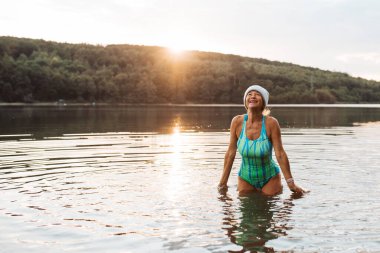 Cold water swimming for elderly women. Senior sporty women standing in lake during cold winter morning, looking at sunrise. clipart