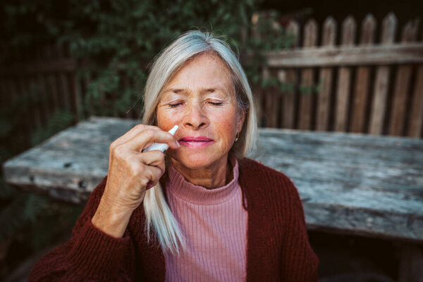 Senior women feeling sick, using nasal spray to help with congestion and stuffiness.