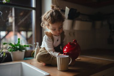 Adorable little girl is pouring tea from the teapot into cup. The sick girl with a cold is sitting on the kitchen counter and concentrating. clipart