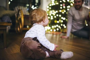 Little baby boy in Christmas home. Cute boy is sitting by Christmas tree enjoying Christmas atmosphere with his father, family. clipart