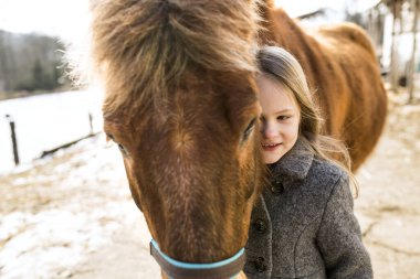 Soğuk kış haftasonunu aile çiftliğinde geçiren, kahverengi tüylü midillisine bakan, ona sarılan genç bir kız.