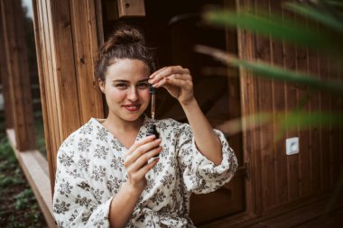 Beautiful woman doing her skin care routine outdoors, wearing bathrobe. Woman using facial skin serum clipart