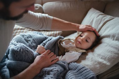 Father taking care of sick girl with fever and cold at home. Daughter lying on sofa, feeling tired clipart