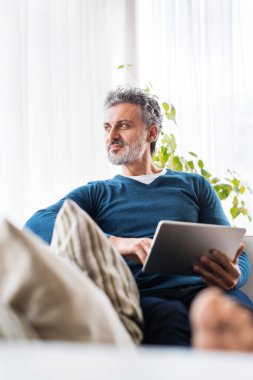Evde orta yaşlı, çekici bir adamın portresi. Tablette TV programı izliyor, oturma odasında kanepede oturuyor, gülümsüyor..