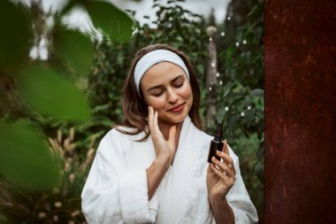 Beautiful woman doing her skin care routine outdoors, wearing bathrobe. Woman using facial skin serum clipart