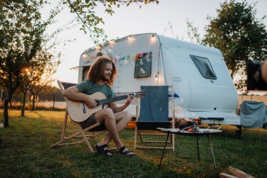 Genç adam doğada bir kamp gezisinde, karavanın önünde oturup gitar çalıyor.