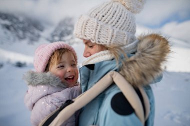 Anne, bebek taşıyıcısında kızıyla karlı bir arazinin ortasında duruyor. Anne ve kız dağlarda kış tatilinin tadını çıkarıyorlar..