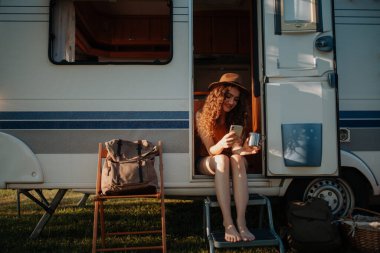 Doğada kamp yapan güzel bir kadının portresi, karavan merdivenlerinde elinde akıllı telefon ve kahveyle oturuyor..