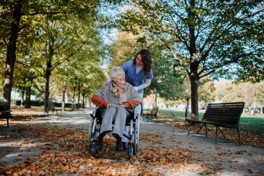 Sıcak sonbahar gününde parkta yürüyen tekerlekli sandalyedeki engelli yaşlı kadın. Genç hemşire tekerlekli sandalye itiyor, yaşlı kadınla konuşuyor..