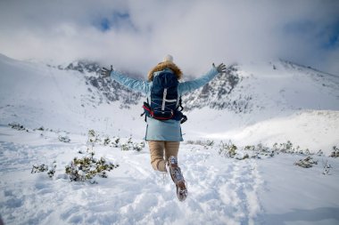 Woman jumping in the middle of snowy nature with open arms, looking at the winter landscape around her. clipart