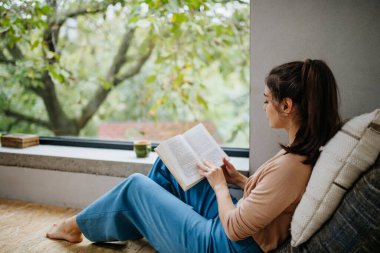 Güzel bir kadın için Hygge anı büyük bir pencerenin yanında elinde bir kitapla oturuyor. Kadın kendine vakit ayırıyor, kitap okuyor..