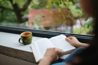 Güzel bir kadın için Hygge anı büyük bir pencerenin yanında elinde bir kitapla oturuyor. Kadın kendine vakit ayırıyor, kitap okuyor..