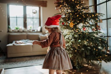 Noel Baba şapkalı şirin bir kızın portresi Noel ağacını Noel süsleriyle süslemeye yardımcı oluyor..