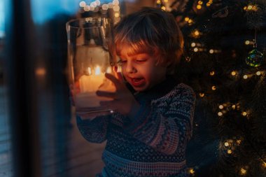 Elinde Noel mumu, arkasında Noel ağacı tutan sevimli küçük çocuk. Evde şenlikli Noel atmosferi.