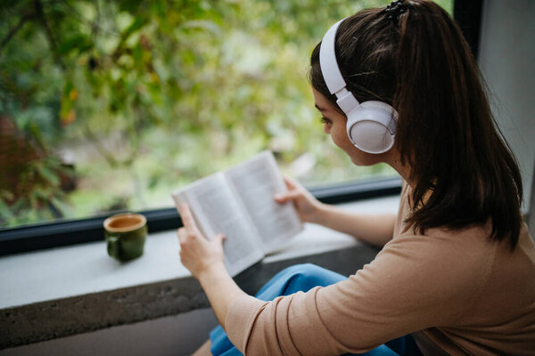Woman sitting by a large window with headphones on her head and book. Woman is enjoying time for herself, reading.