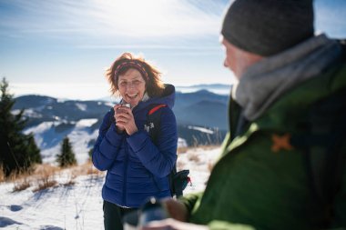 Active seniors resting, drinking warm tea from thermos. Romantic winter hike for elderly couple in snowy mountains. clipart