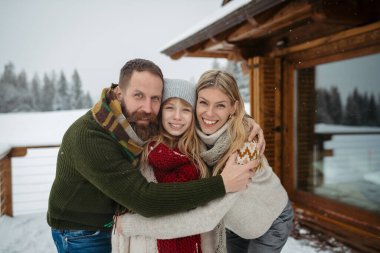 Kar kabininin balkonunda duran aile kameraya bakıyor ve kar yağarken sarılıyor..