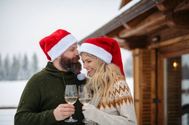 Mature couple wearing Santa hats, standing in front cabin and toasting with wine glasses during slowfall. clipart