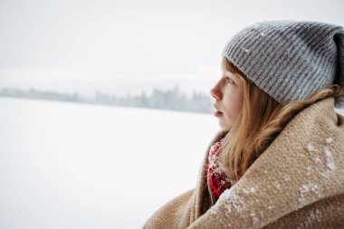 Kalın bir battaniyeye sarılı kız balkonda duruyor ve karlara bakıyor..