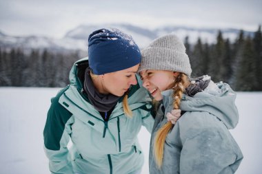 Mom and cute girl hugging, standing in the middle of snowy nature. Hiking in winter mountains. clipart