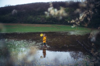 Little girl standing in a shallow marsh with a fishing net, catching frogs. Concept of spring and outdoor fun for child. clipart