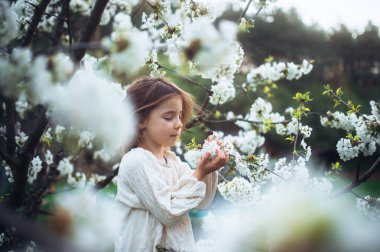 Beautiful young girl holding flowers in her palms, standing in the middle of blooming spring trees. Concept of springtime in nature. clipart