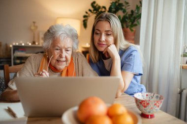 Laptopla çalışmayı öğrenen yaşlı bir kadın. Sosyal hizmetler görevlisi ona yardım ediyor. Yaşlılar için finans ve çevrimiçi bankacılık yönetimi kavramı.