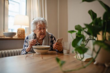 Yaşlı kadın mutfakta oturup çorba içerken akıllı telefon kullanıyor..