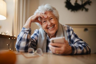 Yaşlı kadın akıllı telefon kullanırken kendine güveniyor, mutfakta oturuyor..