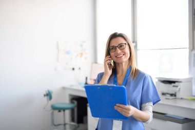 Gülümseyen kadın hemşire modern bir klinikte kendinden emin bir şekilde duruyor ve hastasıyla telefon görüşmesi yapıyor..