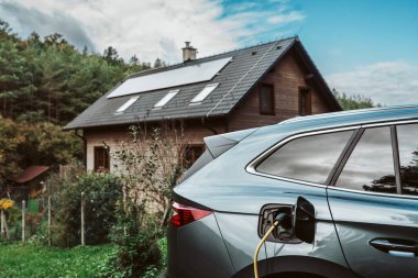 Elektrikli araba evin önünde güneş panelleri çatıda şarj oluyor. Şarj kablosu olan araç.