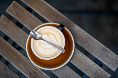 Sıcak bir kafe ortamında, ahşap bir masa üzerinde latte sanatı olan kapuçino bardağının en üst görüntüsü..
