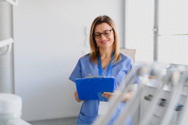 Gülümseyen kadın hemşire, modern bir klinikte güvenle duruyor, randevu çizelgesine bakıyor..