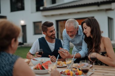 Yaşlı adam, Spring ailesinin barbekü partisinde akıllı telefondan fotoğraf gösteriyor. Aile bahçesi partisi.