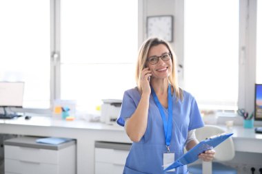 Gülümseyen kadın hemşire modern bir klinikte kendinden emin bir şekilde duruyor ve hastasıyla telefon görüşmesi yapıyor..