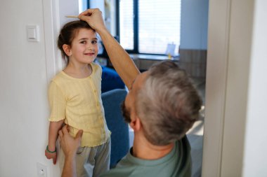 Baba evde kızının boyunu ölçüyor, kapı çerçevesinde sıra yapıyor..