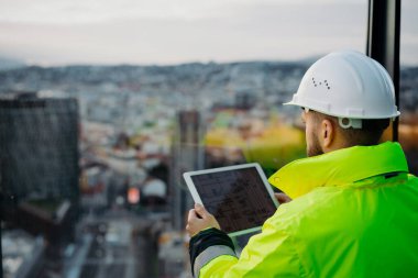 İnşaat alanı yöneticisinin inşaat sürecini denetlemek için dijital tablet kullanan arka görünümü.