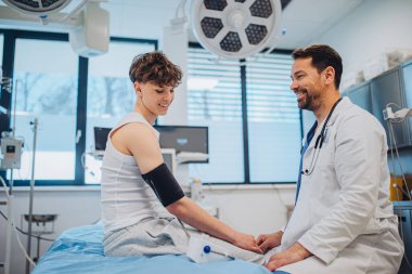 Genç bir çocuk hastane odasında gülümseyen bir doktorla sağlık kontrolünden geçiyor..