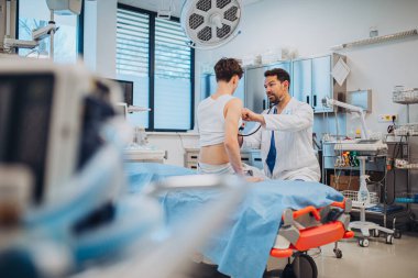 Genç bir çocuk hastane odasında gülümseyen bir doktorla sağlık kontrolünden geçiyor..