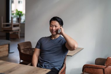 Genç Asyalı adam akıllı telefondan konuşuyor, boş masada oturuyor..