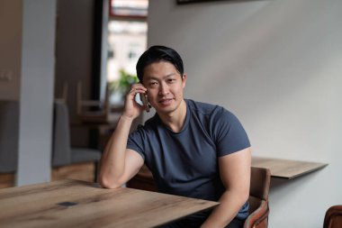 Genç Asyalı adam akıllı telefondan konuşuyor, boş masada oturuyor..